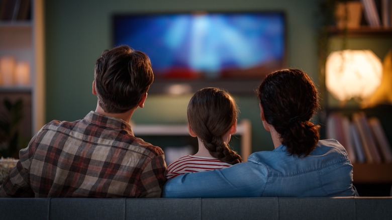 a married couple and daughter watch TV while sitting together on the couch