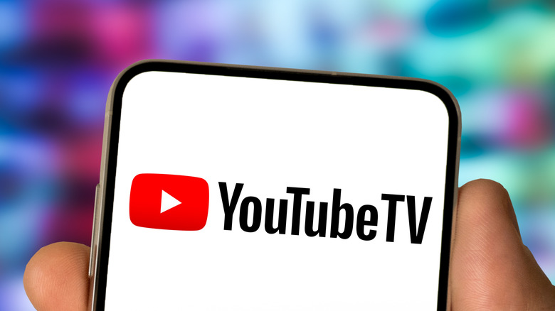 A closeup of a hand holding a smartphone that is displaying the YouTube TV logo, the colorful background is far too blurred to fully make out the details