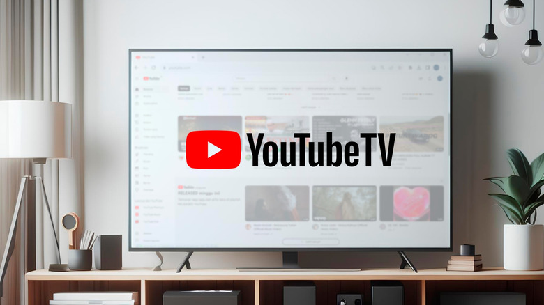 A large flatscreen television displaying the YouTube TV logo sits on top  of a TV stand in what appears to be a modern living room,  the TV is surrounded by a potted plant, a stack of small books, and other assorted items