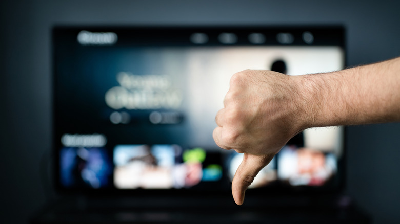 A close-up of hand making the thumbs down sign in front of a TV blurred in the background