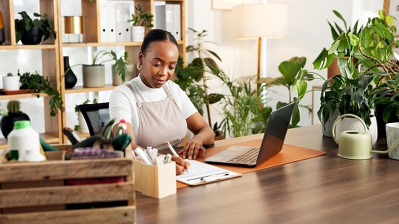 A person at a desk writing on a clipboard while using a laptop