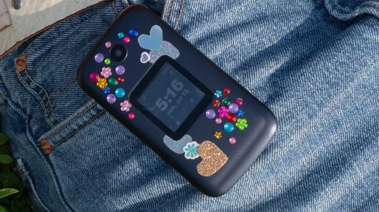 A black Dumbphone 2 covered in colorful stickers sits on top of a user's blue jeans right at the pocket
