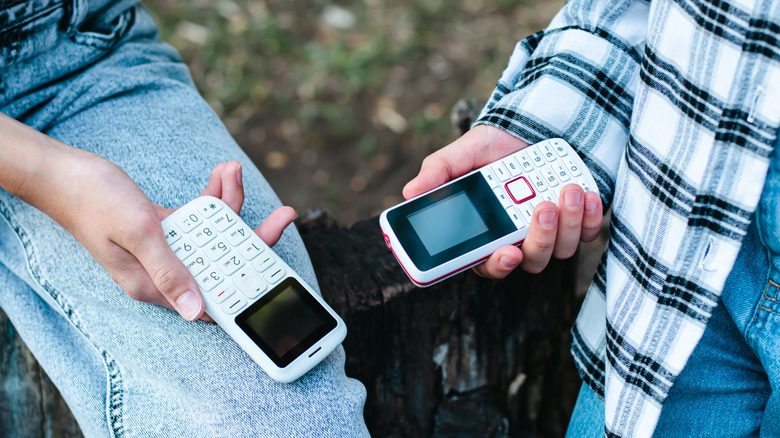 A closeup of two individuals holding "dumb phones" in their hands, both devices are white and small screens