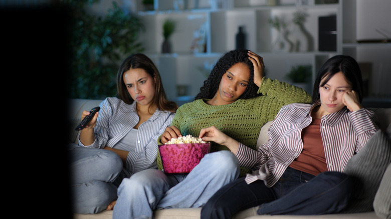 Three friends sitting on couch eating popcorn and looking bored while watching TV