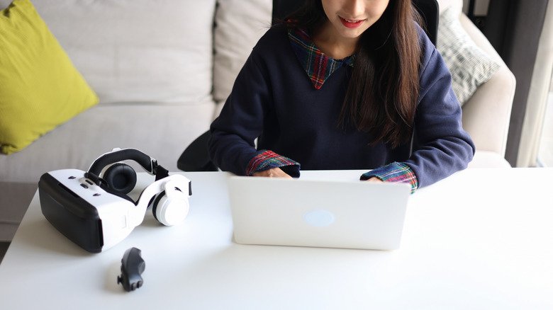 A person using laptop with a VR headset next to them on a white desk