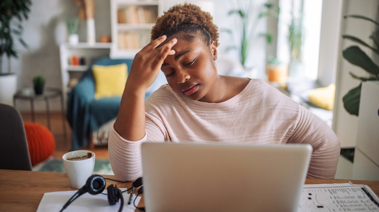 A woman looking annoyed whiled sitting in front of a laptop, with the tip of her fingers touching her forehead.