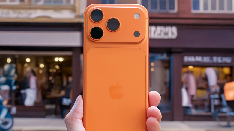 A close-up photograph of a hand holding an orange iPhone 17 on a city street.