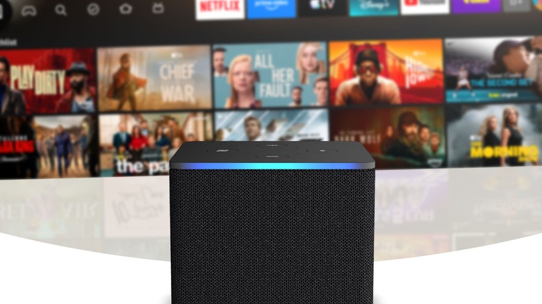 A close-up image of the Amazon Fire TV Cube with a TV in the background displaying the Fire TV interface