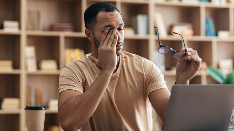 A man taking off his glasses to rub tired eyes while sitting in front of a computer