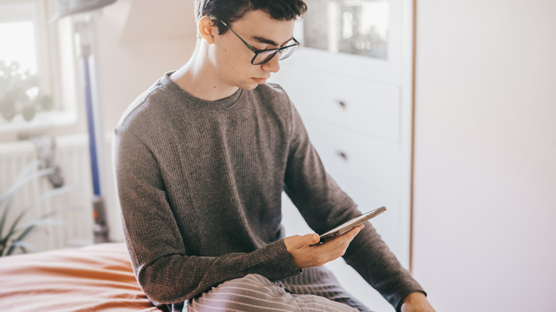 A young man in glasses sitting on a bed looking at a smartphone