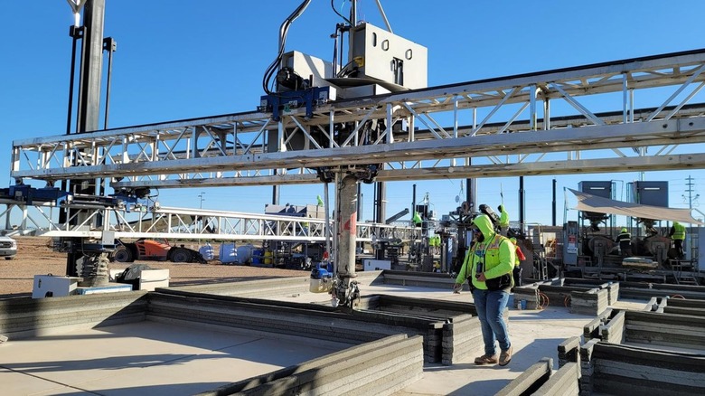 A construction worker observing a 3D printer arm lay down concrete for a 3D-printed house
