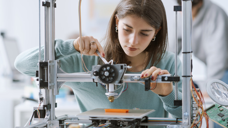 Woman adjusting a 3D printer while it creates a small object on the print bed.