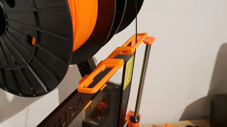 Orange 3D printed filament guide attached below a spool on a 3D printer.