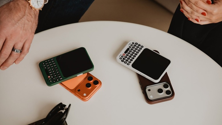 Two Clicks Communicator smartphones with QWERTY keyboards placed on top of iPhone 17 Pro models on a table.