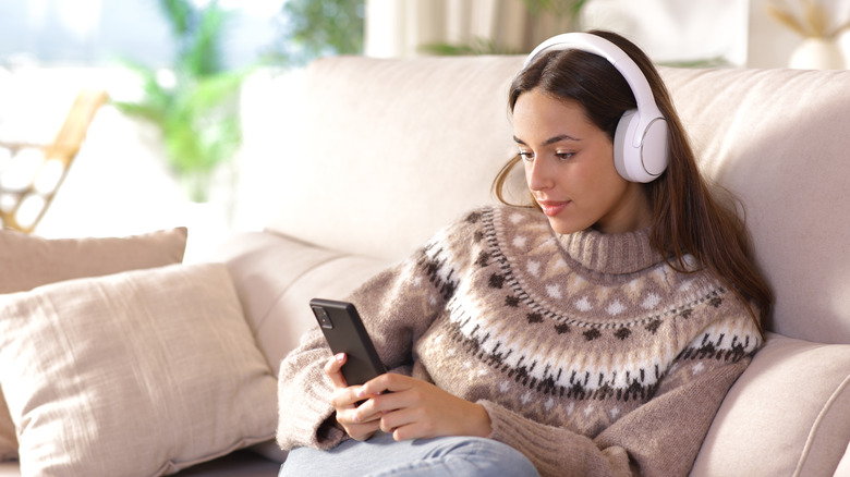 Woman in winter listening to audio using phone and headphones sitting on a couch at home