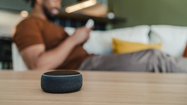 A picture of the Amazon Echo Dot Bluetooth speaker on a table, with a man sitting on the couch while on his phone