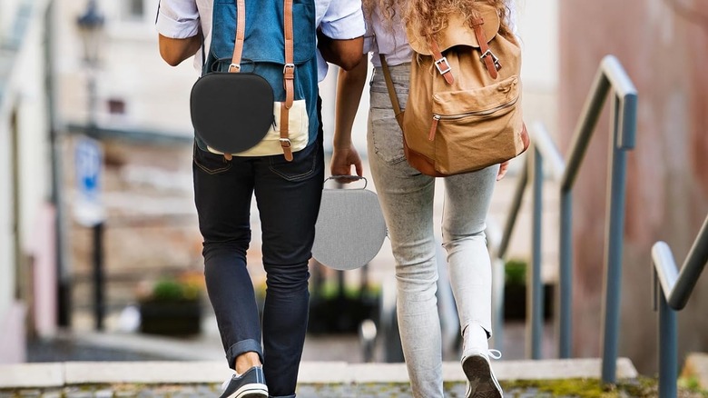 Two people seen from behind walking carrying backpacks. Both have AirPods Max cases, one attached to a backpack and one being carried.