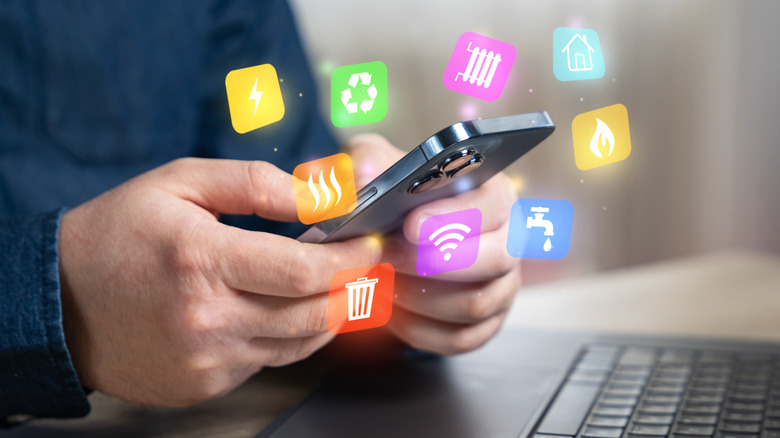 Two hands holding an iPhone while the individual is seated in front of a computer, icons representing various monthly bills are super imposed around the iPhone