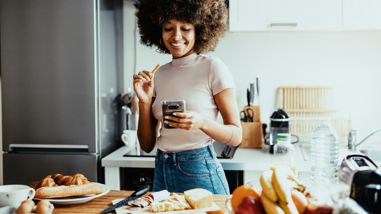 A woman standing in a kitchen surrounded by bread, eggs, and a fruit bowl, she is holding a cellphone in her left hand and a small bit of food in her right