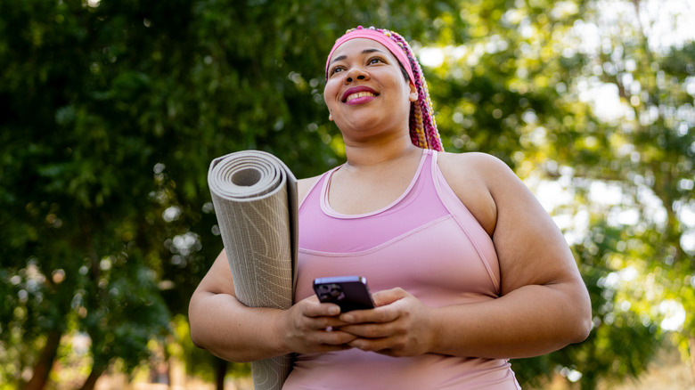 A woman standing outside holding a cellphone with both hands, she has a yoga mat under her right arm as is standing in front of trees