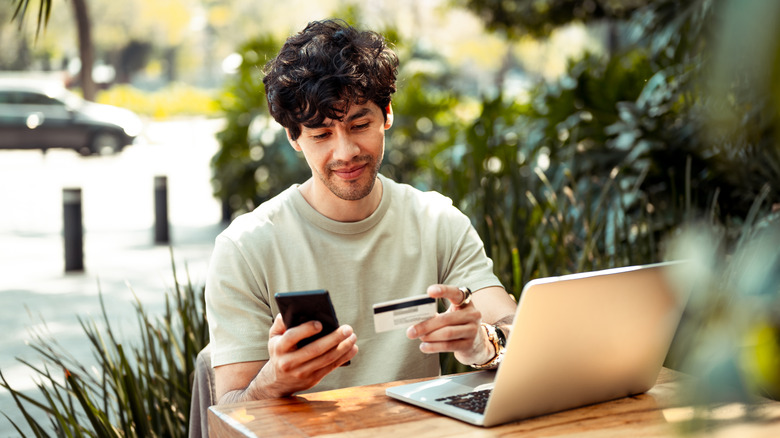 A man in a light green shirt sitting outside as he holds a cellphone in one hand and a credit card in another, he is seated in front of a laptop