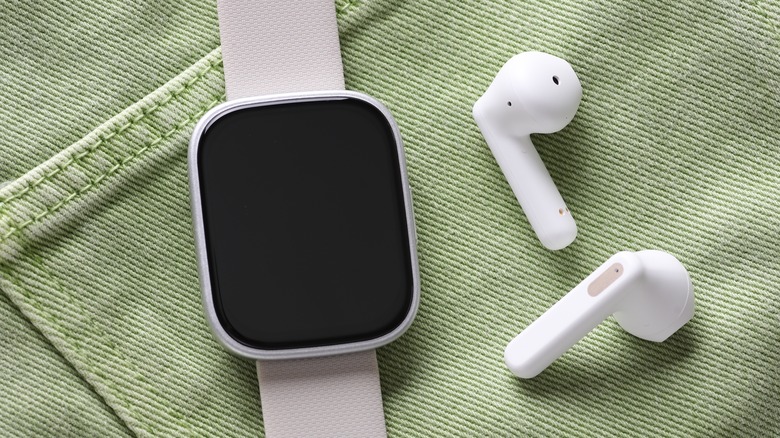 A smartwatch with its screen sleeping laying on a pair of green jeans next to two wireless earbuds