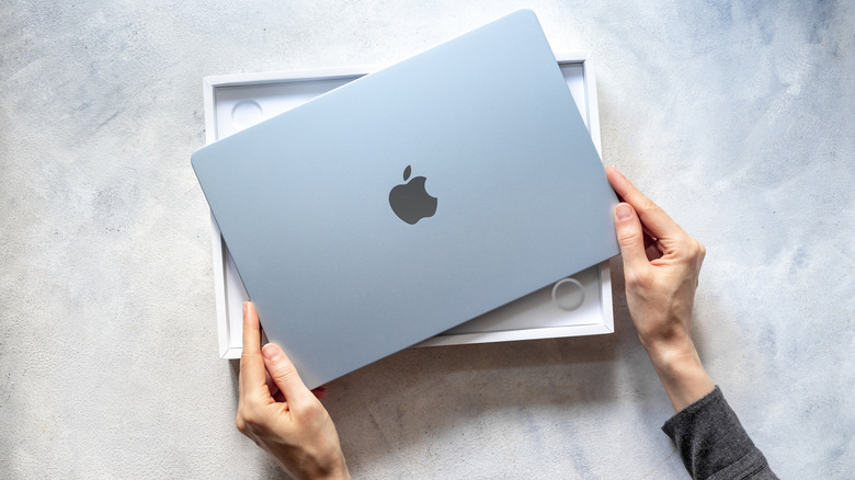Two hands on the corners of a MacBook that is sitting atop its box, both items on a grey counter