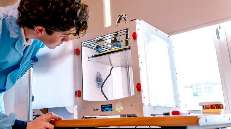 Person inspecting enclosed 3D printer with transparent protective chamber door open