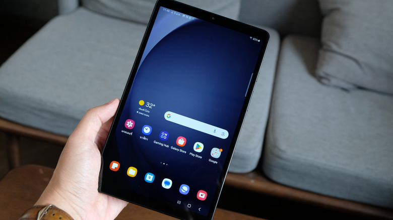 A hand holding a Samsung tablet in front of a grey couch