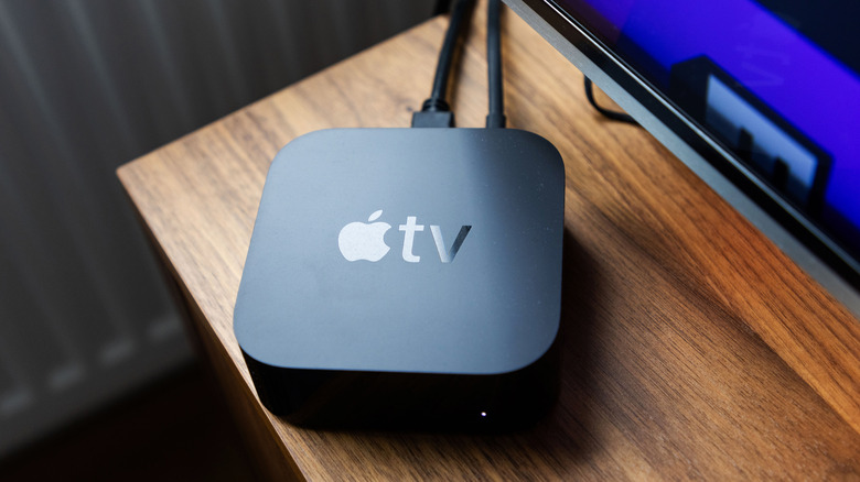 A black Apple TV sitting on the corner of a television stand, the corner of the TV somewhat visible in the picture
