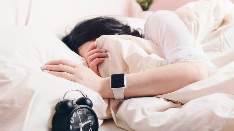 An individual covers their face as they lay in bed, an alarm clock and an Apple Watch with a with a white wrist band are prominent in the photo.
