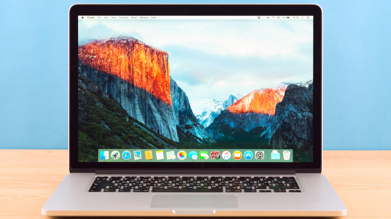 A MacBook sitting on a wooden table against a blue background, the Mac is displaying its desktop, which has a background containing mountains