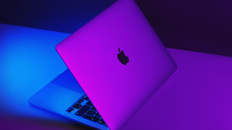 A MacBook sitting on a table, the machine partially opened, purple light washes the backside of the device, whereas blue light is prominent toward the front of the device