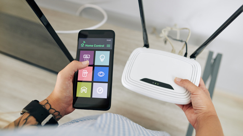 person holding a smartphone in one hand and a router in the other, trying to connect a thermostat to the network