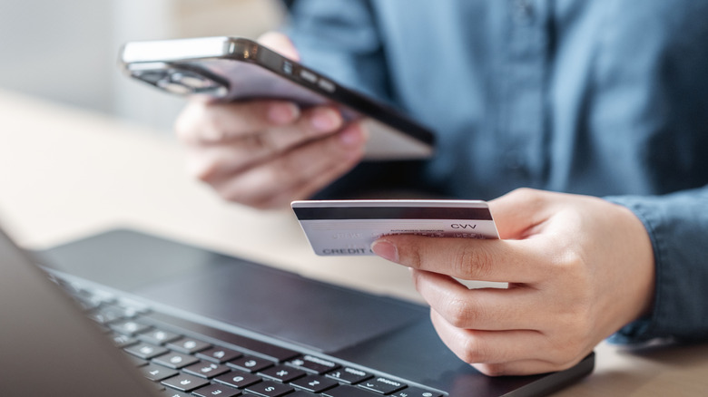 person holding a smartphone in one hand, credit card in the other, with a laptop placed in the front