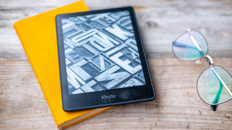 A Kindle eReader on a notebook sitting on a wooden table, a pair of glasses sit next to it