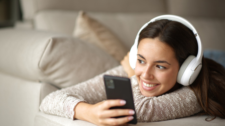 A woman smiles while wearing headphones and lying on her couch, she holds her phone in hand