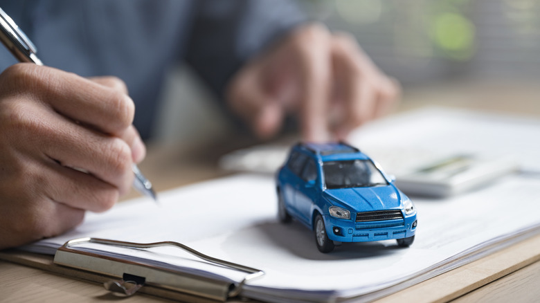 A pair of hands on a desk, with the right hand holding a pen, a clipboard with papers sits atop the desk, as well as a small, blue toy car