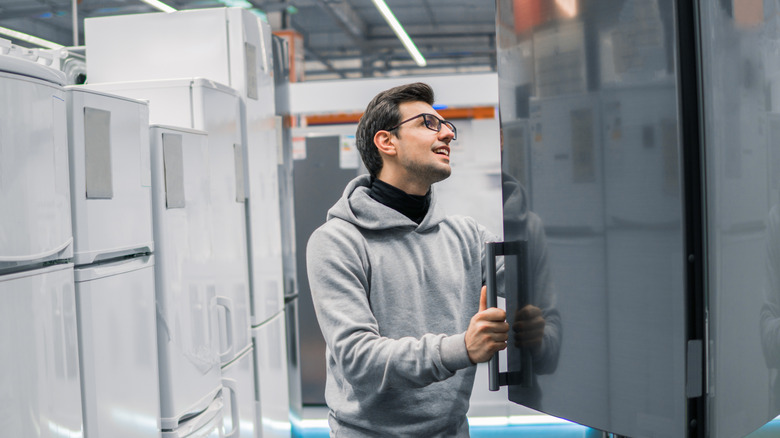 A man looking inside an open refrigerator in a store aisle filled with white, unmarked fridges