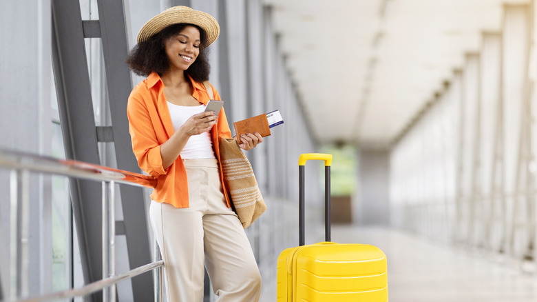 A woman smiling at her phone beside a yellow suitcase in an airport walkway