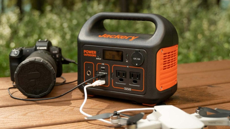 A black Jackery Explorer 300 Plus portable power station charging a drone and a camera on a brown table