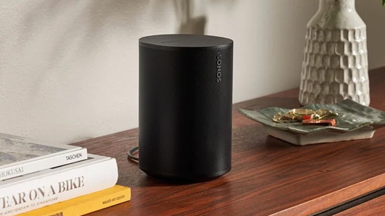 A black Sonos Era 100 speaker placed on a brown table beside books and trinkets