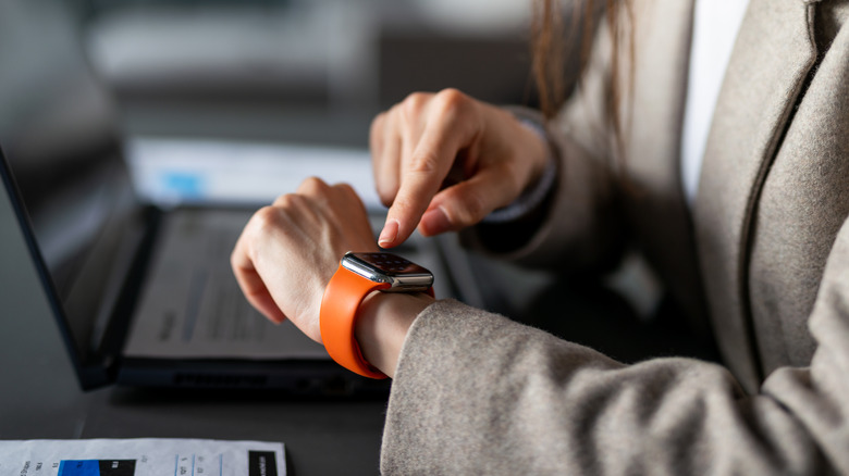 A woman using a smartwatch while working on a laptop.
