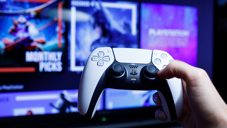 A hand holding and playing a PlayStation 5 controller with the TV in the background