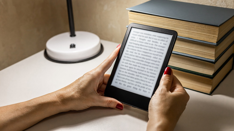 A person using a e-reader near a table lamp in front of some books on a wooden desk