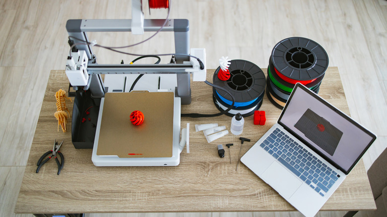 3D printer workstation with filament spools, tools, printed parts, and a laptop on a desk.