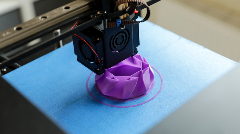 3D printer nozzle creating a purple decorative object on a blue print bed.