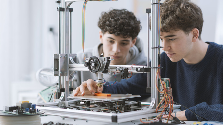 Two people closely inspecting a 3D printer as it prints a small orange object.