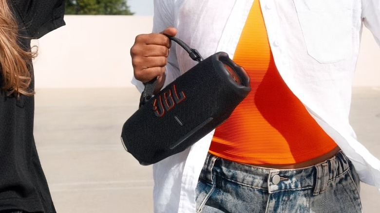 A closeup of an individual dressed in white and orange holding the JBL Charge 6 in their right hand