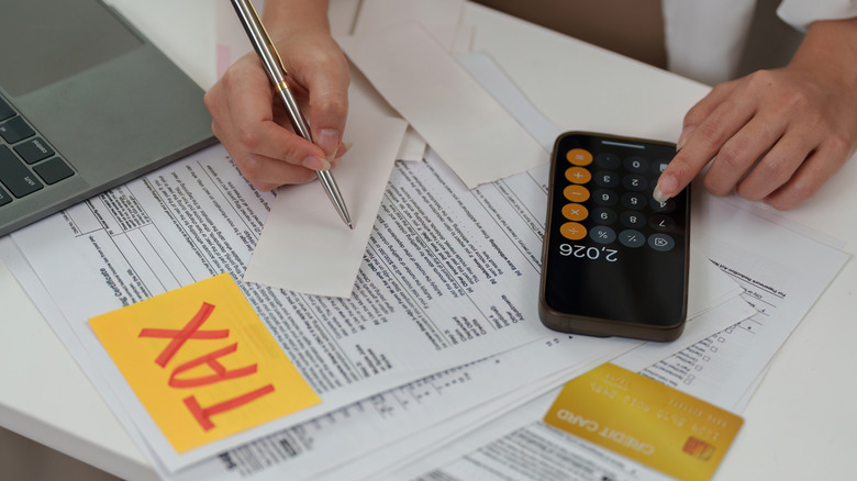 A hand on a calculator app with tax forms in front of a laptop.